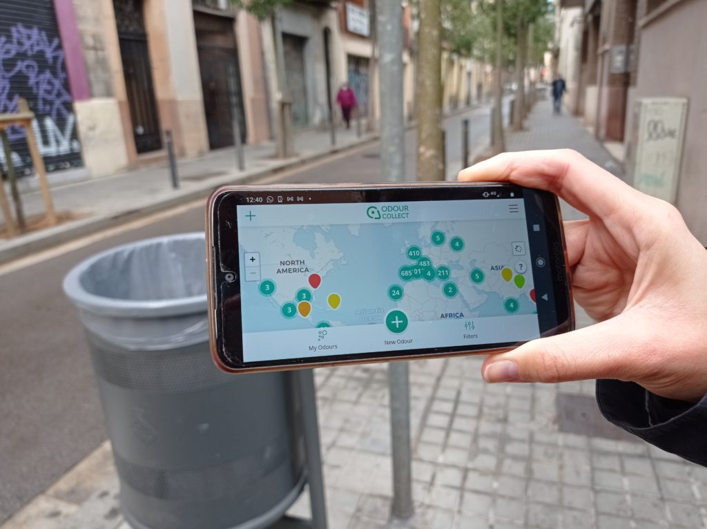 The OdourCollect app shown on a smarphone held by a human hand with a street in the background
