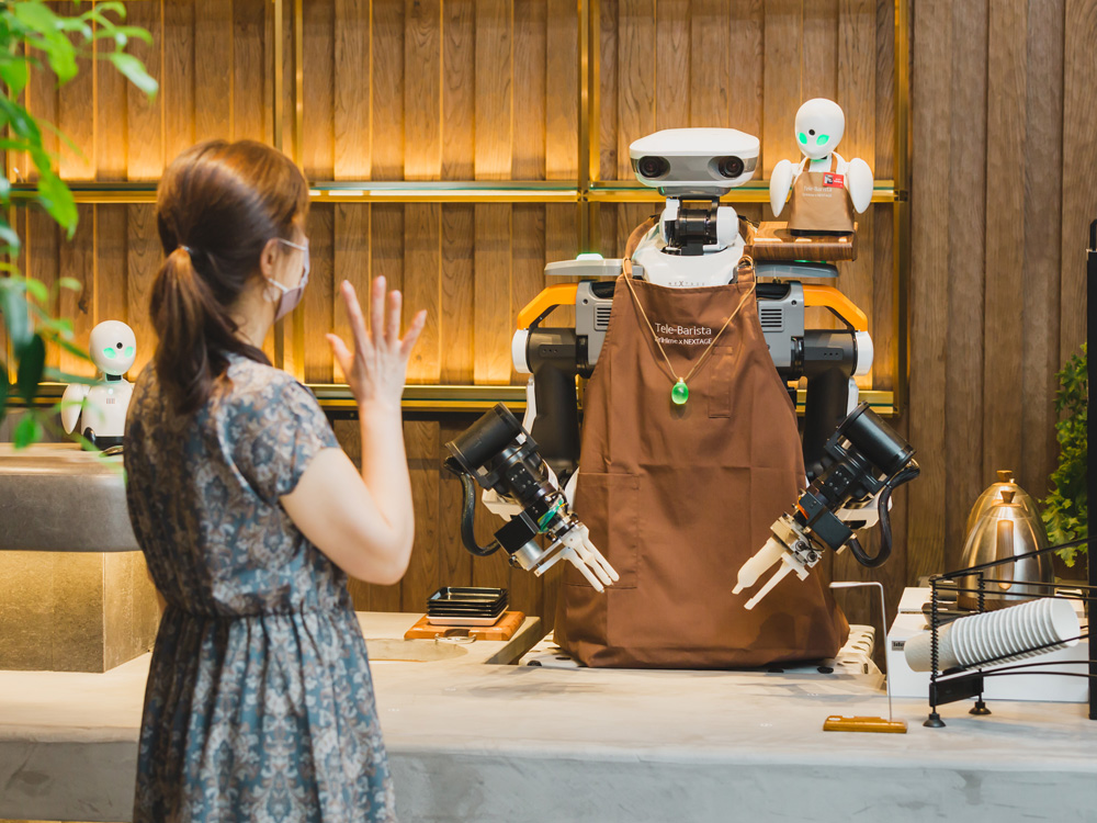A girl wearing a dress and a ponytail waives at the robot humanoid telebarista at the coffee bar