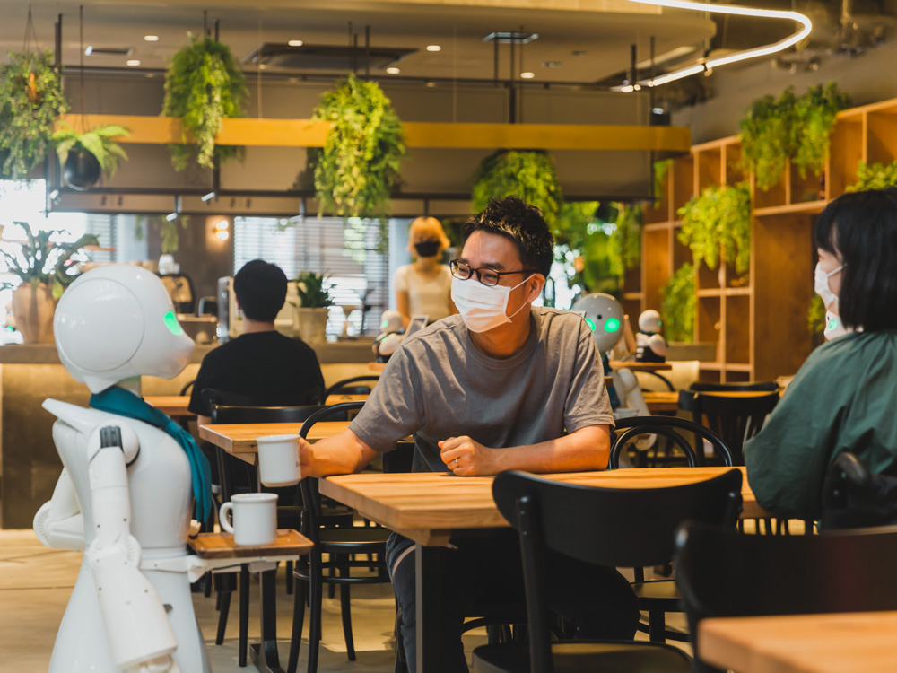 DAWN cafe avatar robot serves coffee mug to human customer sitting down and wearing a face mask and glasses
