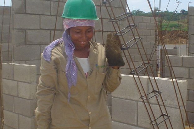 A woman wearing a builder hat and construction gloves smiles in a construction site