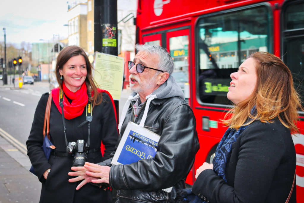 BEarded man with glasses stands outside a double decker bus with two tourists holding a guide